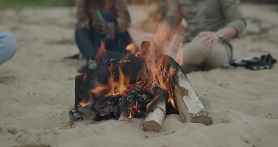 Wood fire burning on sand and two people in the background with green bottles in their hands