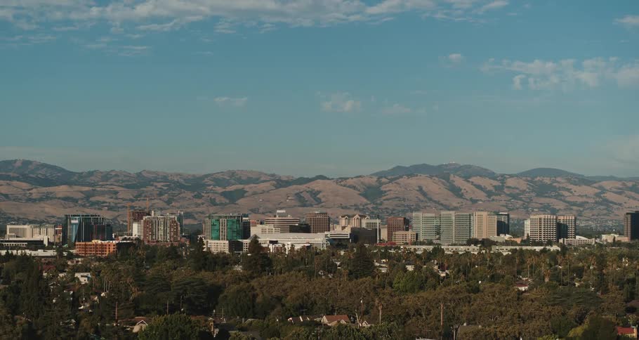 A city with buildings with trees in the foreground with a mountain range the sky and clouds in the background