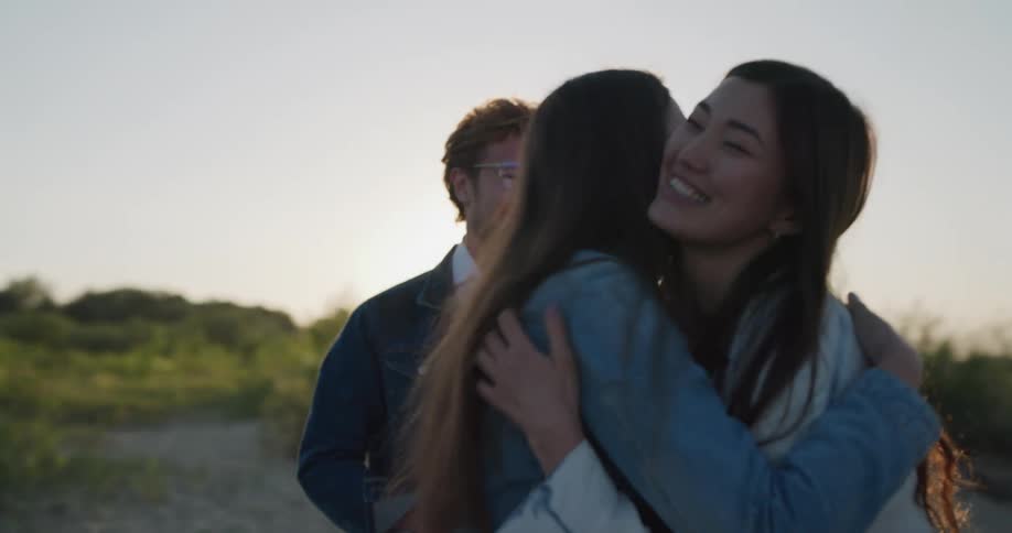 Two men and a woman walk along the beach talking and laughing and then meet two other women and they all hug