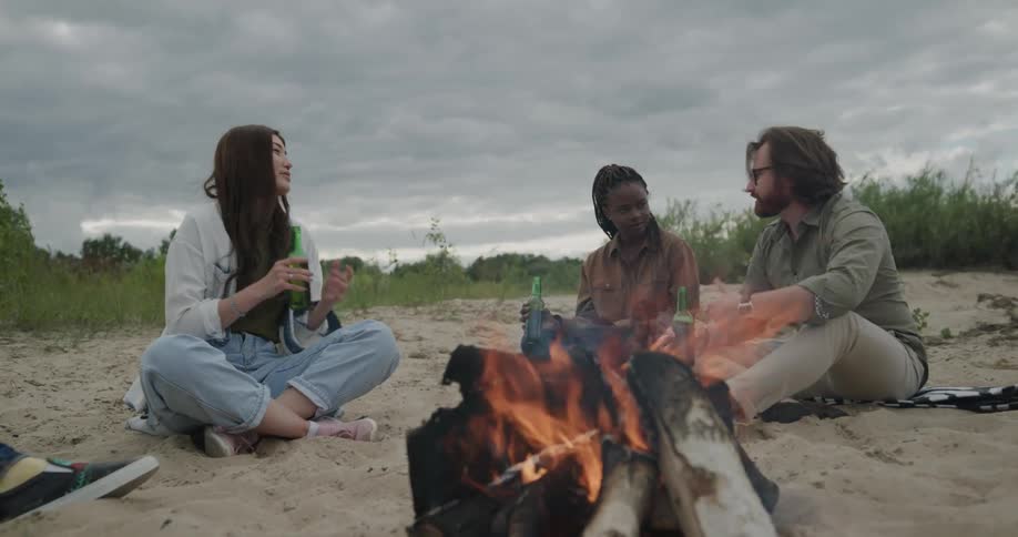 Two women and a man sit around a fire on a beach and talk and drink beer