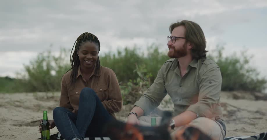 A man and a woman sitting on a shore with beer bottles with a fire in the foreground and the sky and clouds in the background