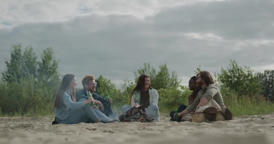 Two men and three women sitting around a bonfire drinking beer with trees the sky and clouds in the background
