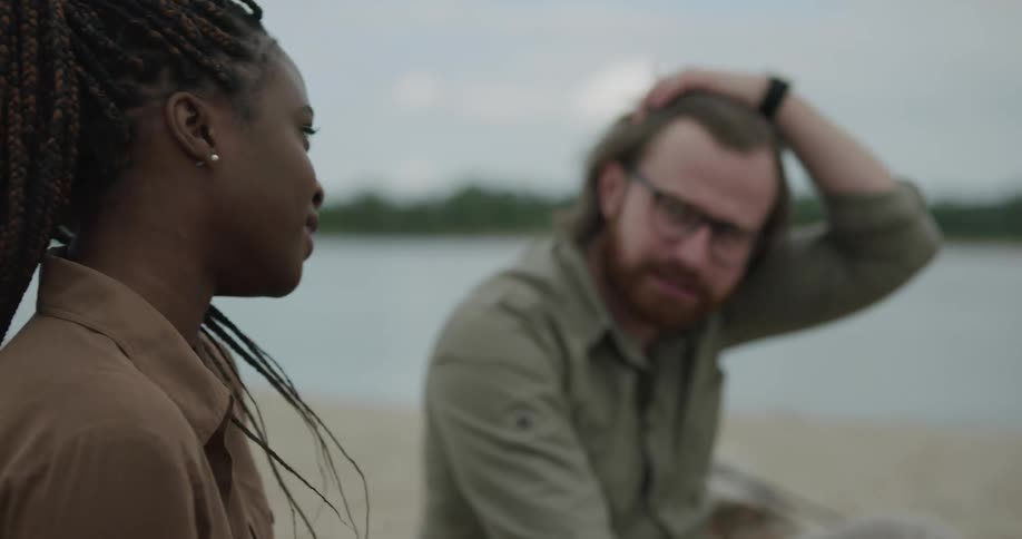 A man and a woman sitting on a shore talking with trees the sky and clouds in the background