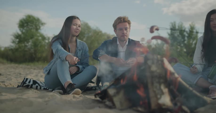 A man and two women sitting by a bonfire roasting marshmallows and sausages with trees the sky and clouds in the background