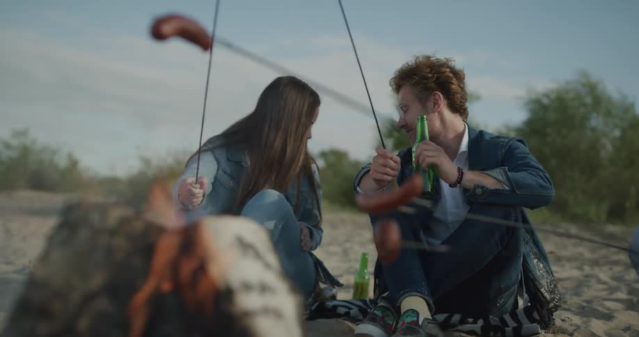 A man and a woman sitting in front of a bonfire roasting sausages and drinking beer with trees the sky and clouds in the background
