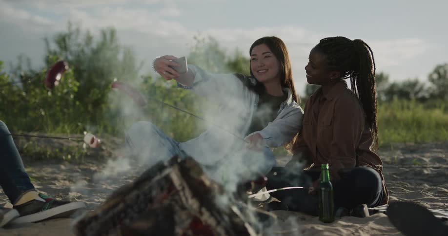 Two woman sitting by a bonfire roasting sausages and taking pictures of themselves with a smart phone with trees the sky and clouds in the background