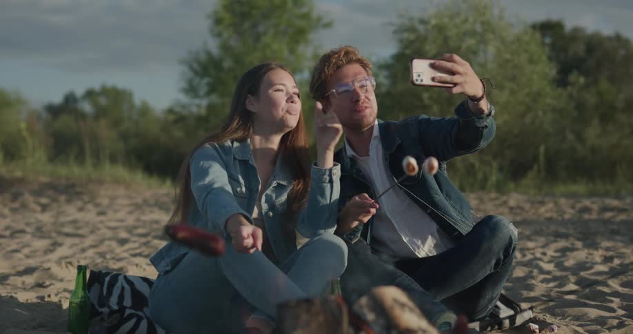 Group of friends around a bonfire taking selfies on the smartphone and cooking sausages on the fire