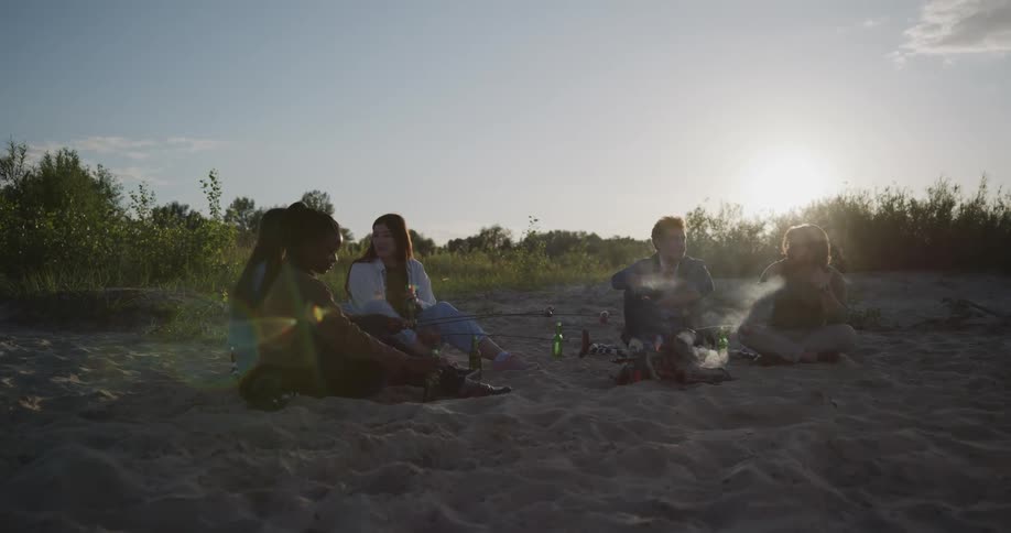 A group of friends sit around a fire on a beach and cook hotdogs and drink beer as they talk