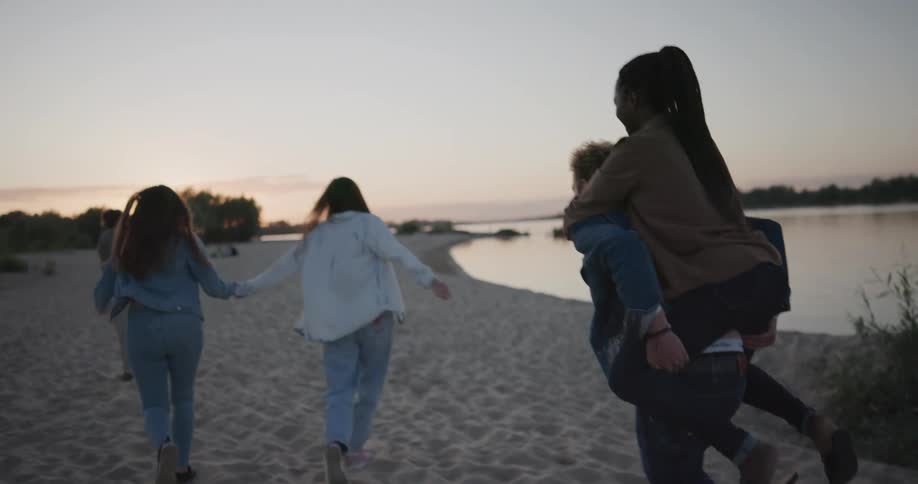 Man carrying woman piggyback hurries to keep up with friends as they walk along sandy shore toward red tinted horizon