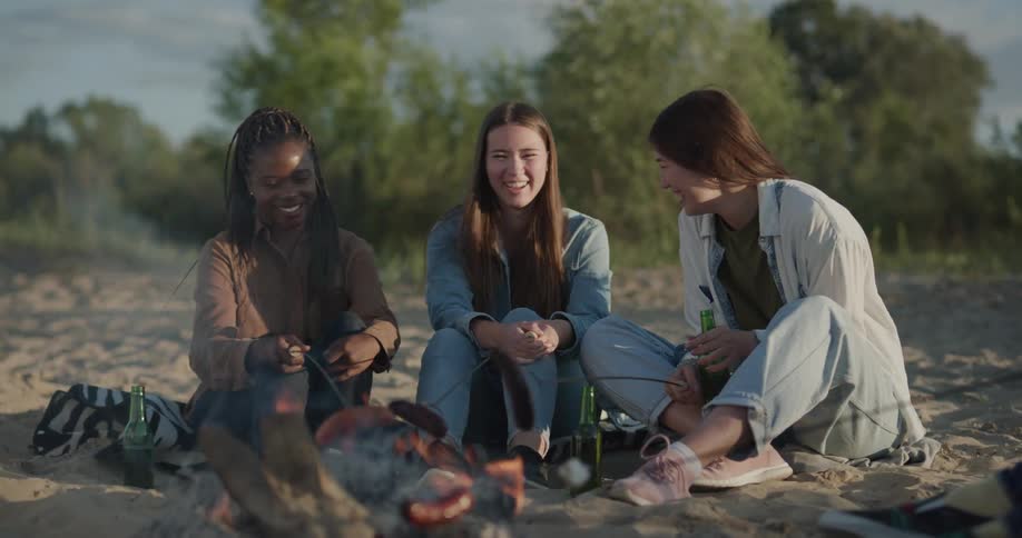 Friends sit around campfire roasting franks and marshmallows