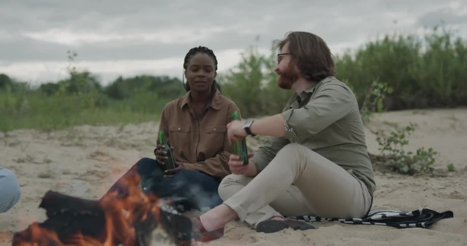 A man and a woman sitting in front of a bonfire with trees the sky and clouds in the background