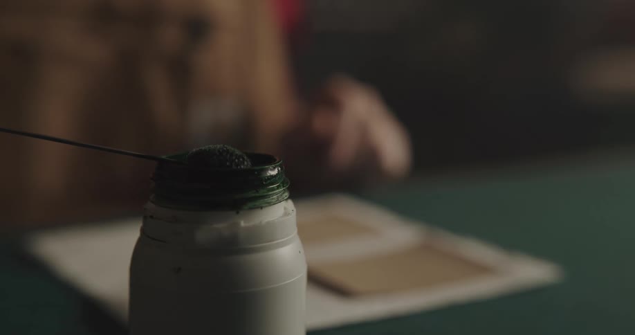 A man dips a round brush in a jar of paint and then paints a square of paper