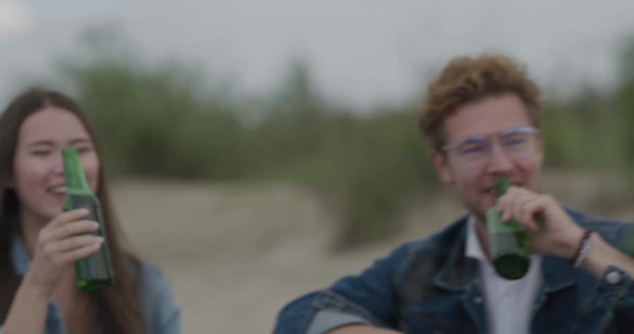 A group of friends sit on a beach and knock their beer glasses together and then take a drink