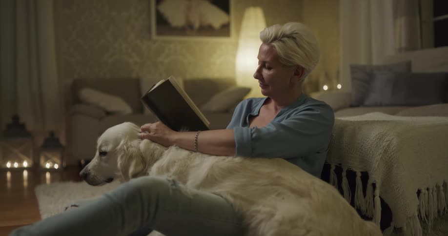 White woman sitting at the floor petting her dog while reading a book