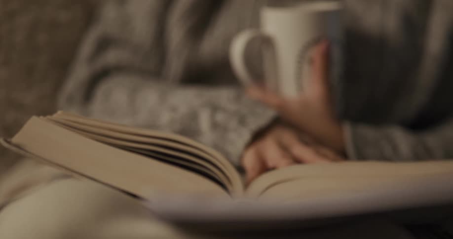 Woman wearing gray sweater is holding a coffee cup in one hand as she turns the pages of a book that is sitting in her lap with the other hand