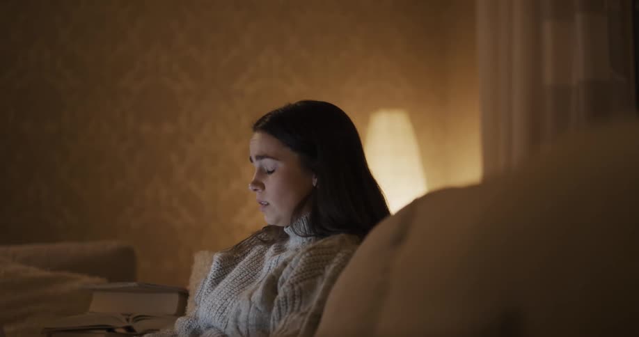 A woman in a room sitting on a couch with a wall and a lamp in the background