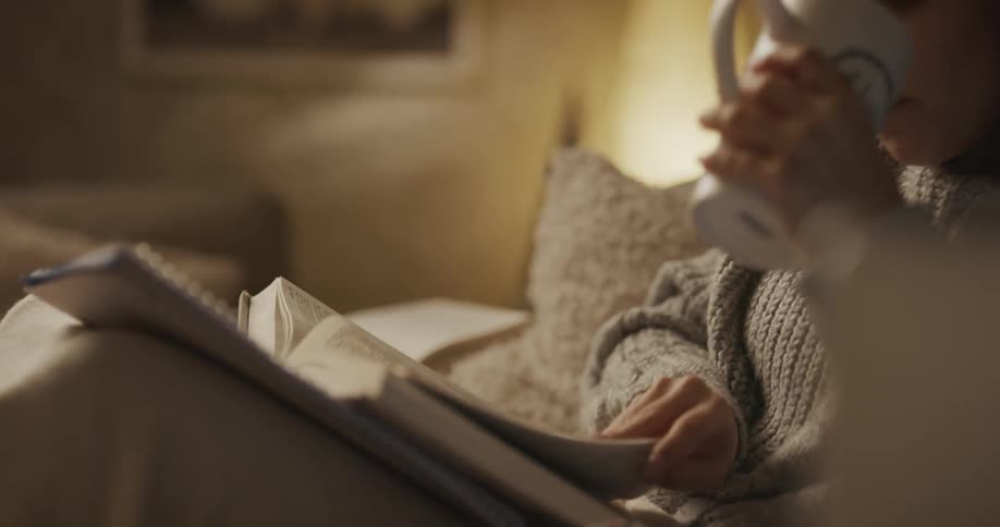 A woman in a room sitting on a couch reading a book and drinking from a mug with a lamp in the background