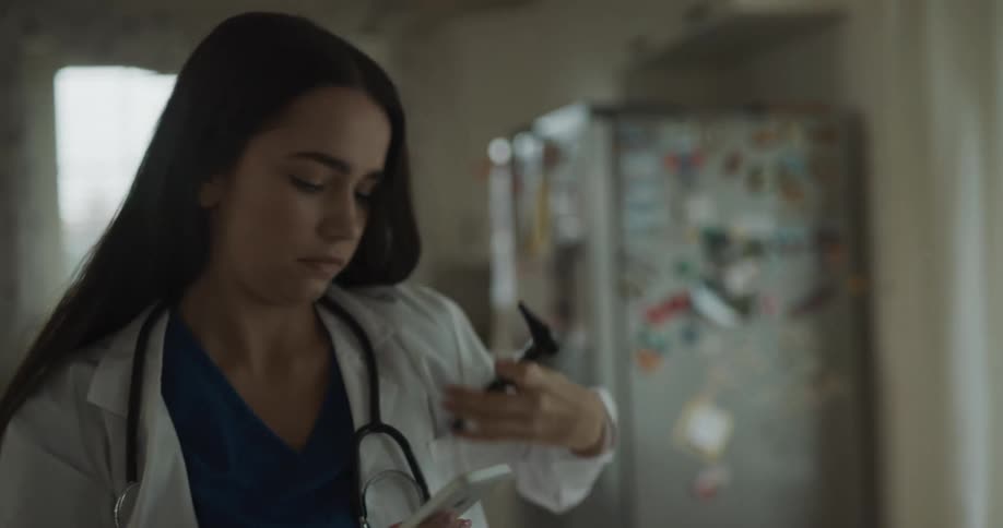 A woman with a lab coat and a stethoscope looking at a smartphone with a fridge and a window in the background