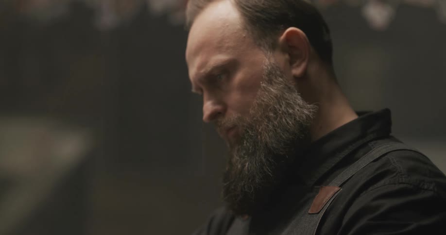 Barber with black shirt apron and beard cuts mans hair in barbershop
