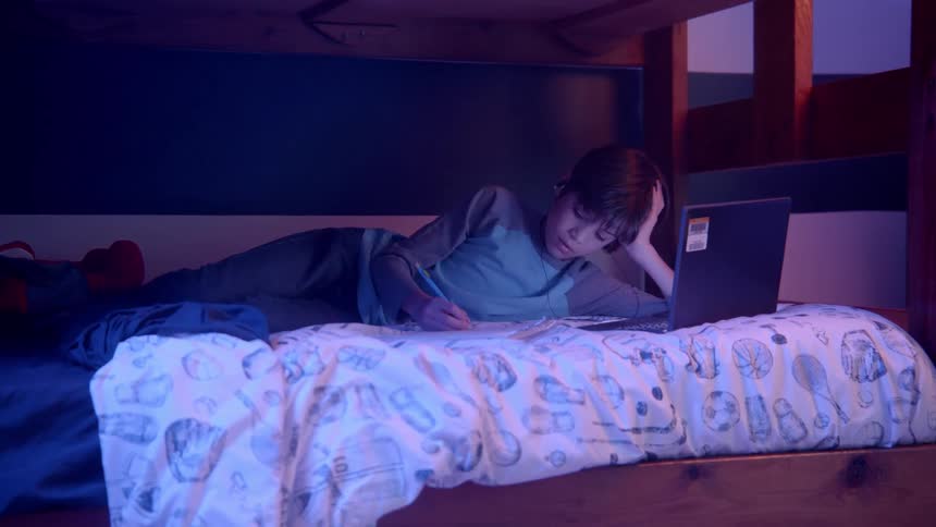 A boy in a bedroom laying on a bed looking at a laptop and writing in a book