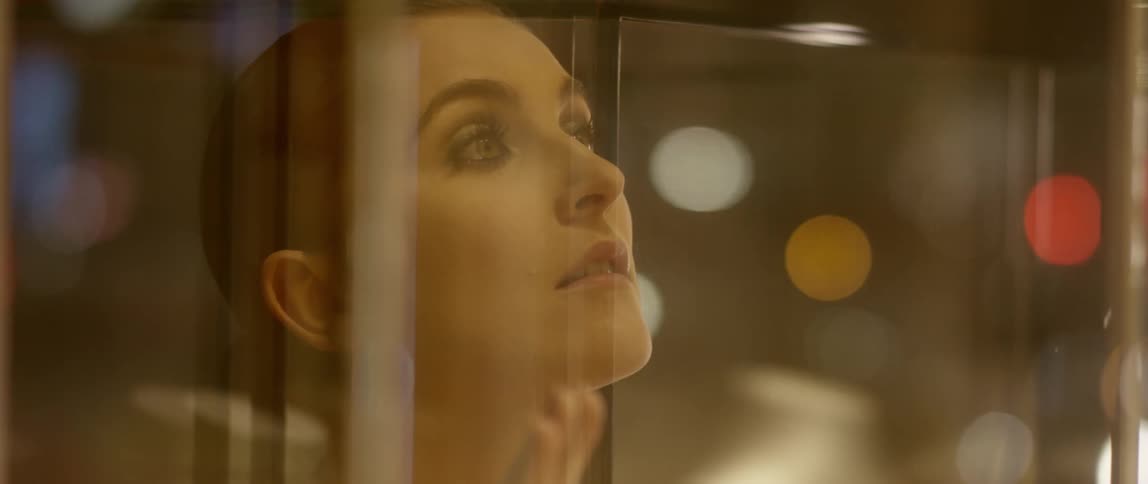 A woman with a necklace and a red dress walking up and touching a window with lights reflecting off of it