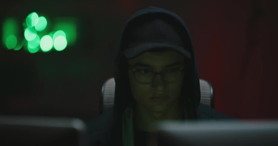 A teenager adjusts his hat while sitting in front of two computer monitors with a black hoodie