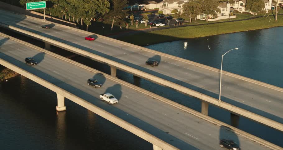 A highway bridge over a river with vehicles driving on it with trees and houses in the background