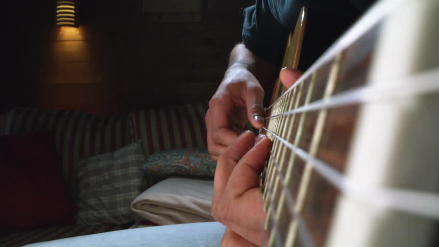 Extreme close up of an acoustic guitar neck being played by a man sitting in a room at home
