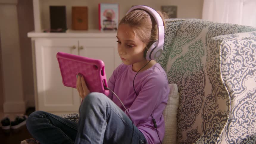 White girl sitting at armchair using her tablet while listening to music on her headphone