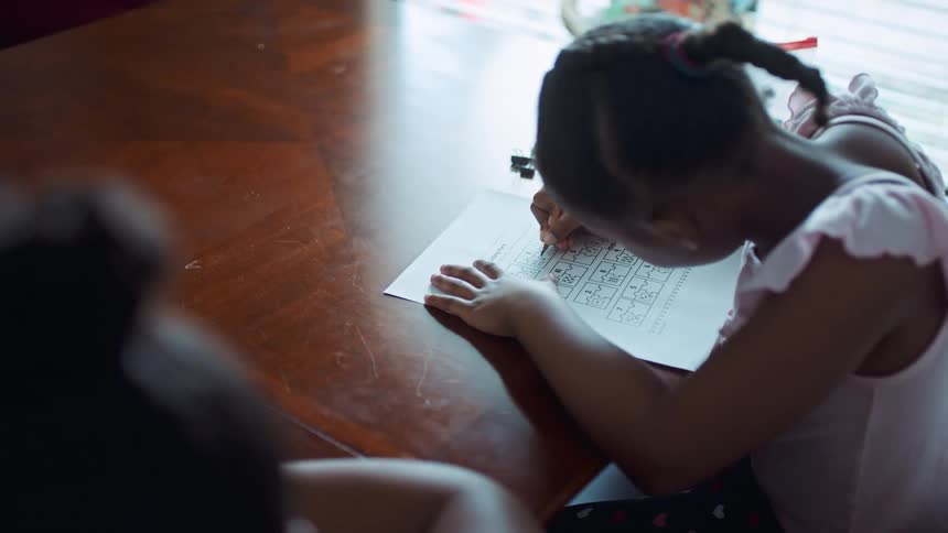 Two young sisters write on worksheets while doing homework together