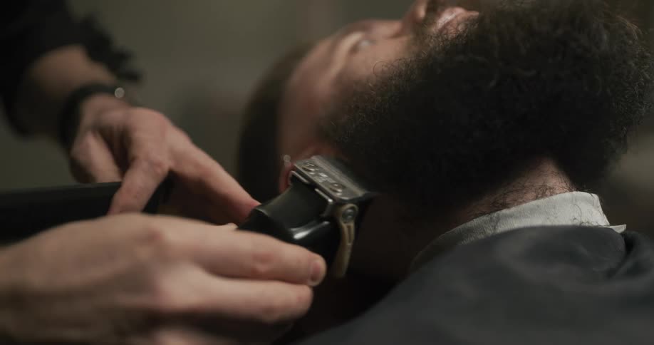 A man in a barbershop shaving another mans beard with electric shavers and a comb
