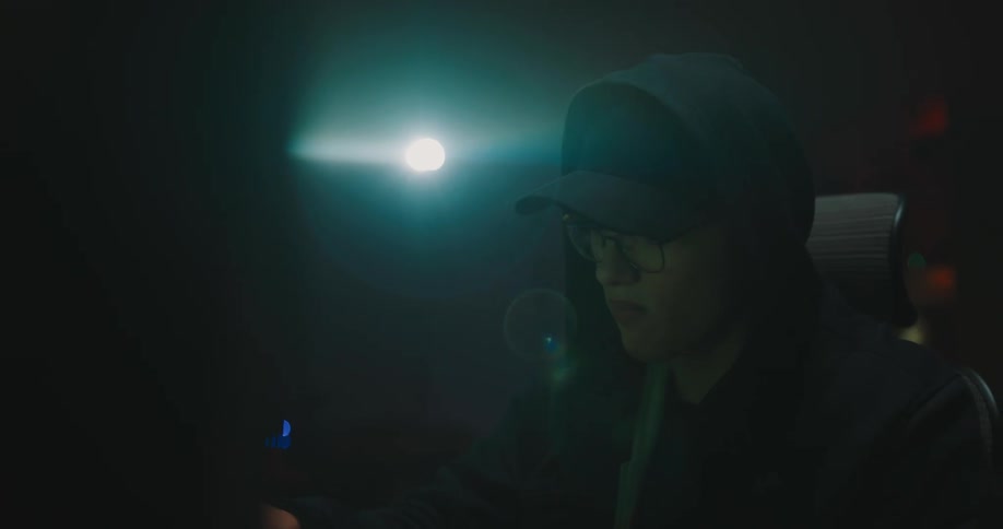 A man with a hat and glasses sitting on a chair at a desk in front of a computer and typing on a keyboard