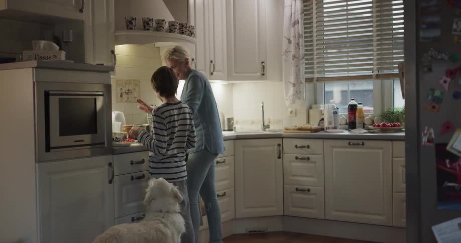 A young boy and his dog run in to a kitchen where his mom is talking and they talk and she sends him away