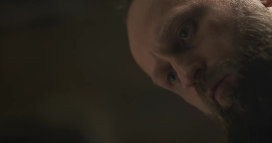 A large bearded man stares intently at the floor