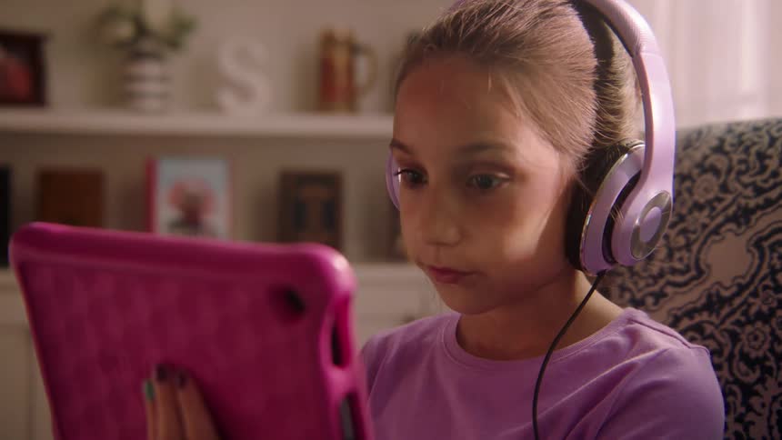 A girl sitting on a cushioned chair in the living room of a house while wearing headphones nods as she watches the screen of the tablet she is holding