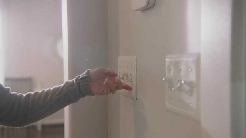 A woman walks through her home as flips up light switches in a hallway as she passes