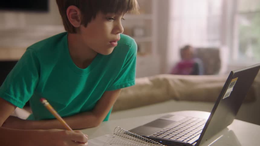 Boy studying on his laptop and writing down on his notebook at living room
