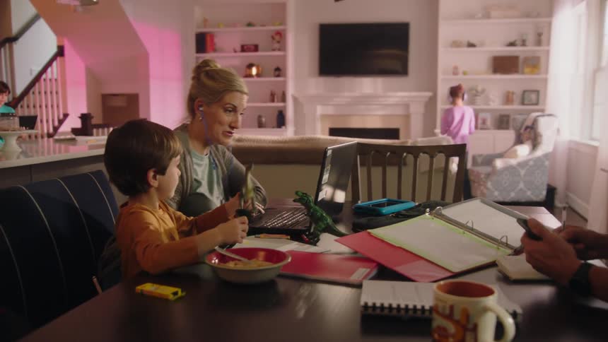 A woman works on her laptop as her son plays with toys next to her and her husband gets up to answer his phone