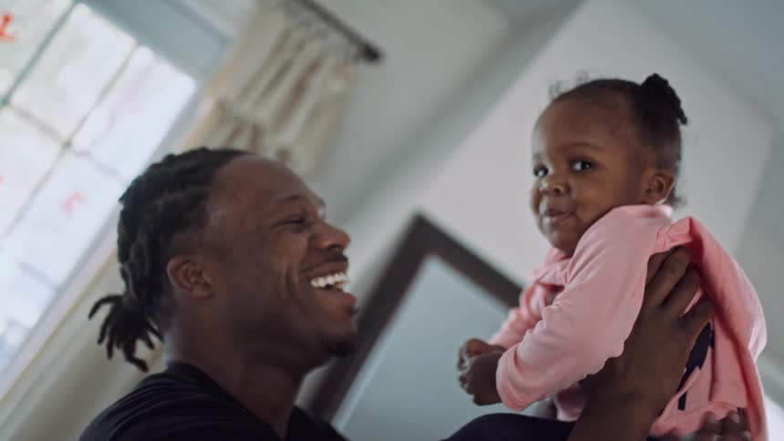 A young girl looks into the camera while being held into the air by a laughing man in the living room of a house during the day