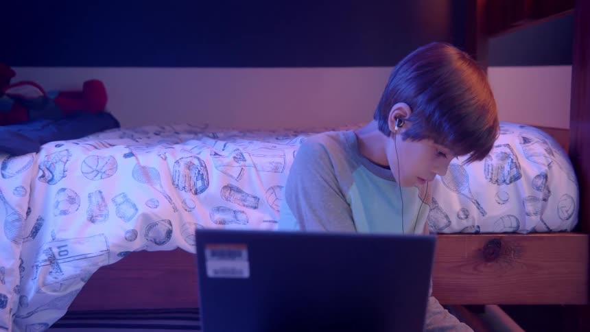 Boy with grey pyjamas and earphones uses laptop and writes in a notebook in his room