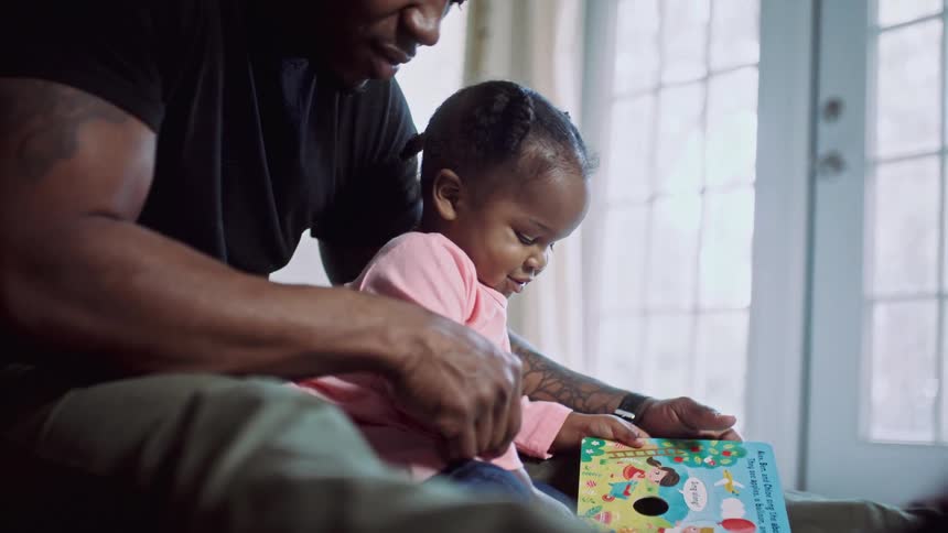 Father and daughter sitting together at the floor inside the house while he helps her read a children book