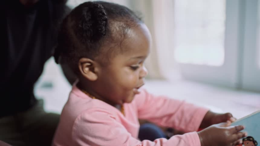 A young girl opens a picture book while sitting in a mans lap during the day