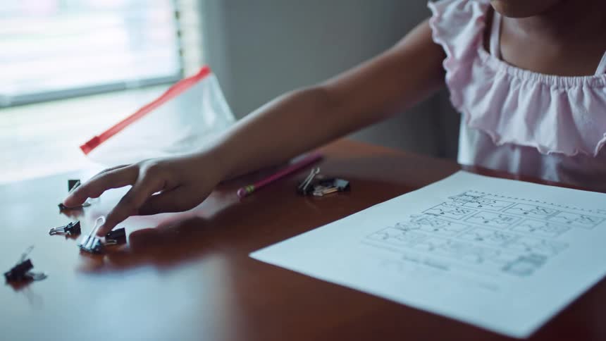 A little girl in a room sitting on a chair at a table with a wall and a window in the background