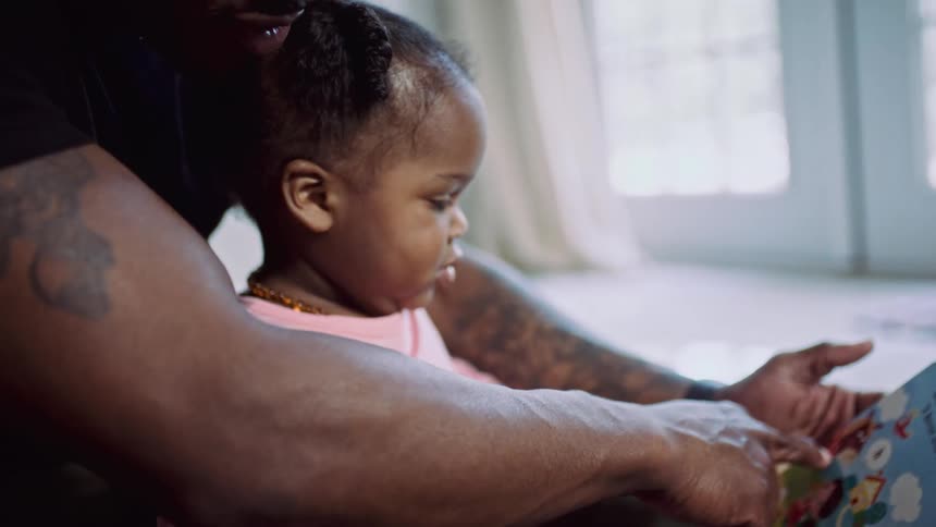 A dad sits with his daughter in his lap on the floor reading a book