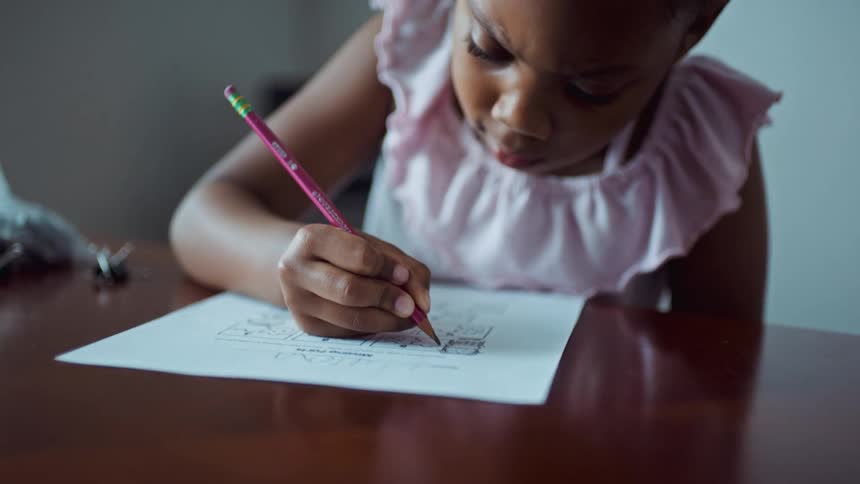 A girl sitting at a table drawing on a paper with a pencil with a wall in the background