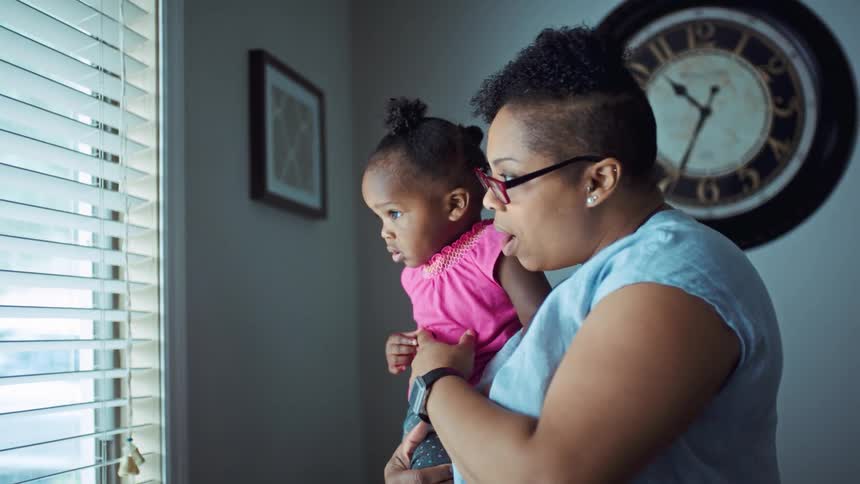Mother holding her baby daughter on her hands looking out the window at home