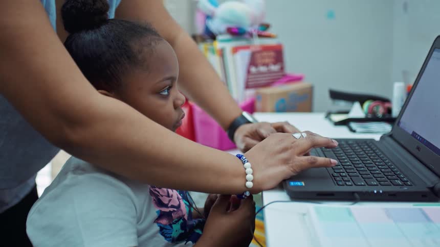 A woman in a room helping a girl with a laptop with a wall and a window in the background