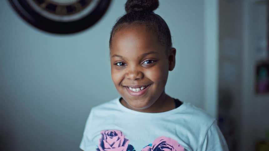 A young girl poses in her home in a top bun and flowered shirt