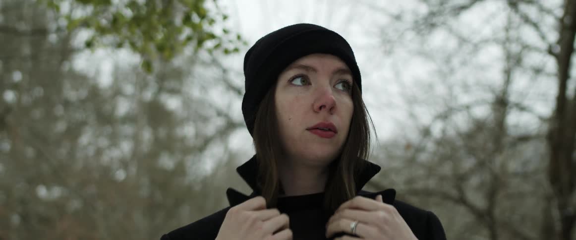 A woman holds her coat collar around her neck while walking through a forest