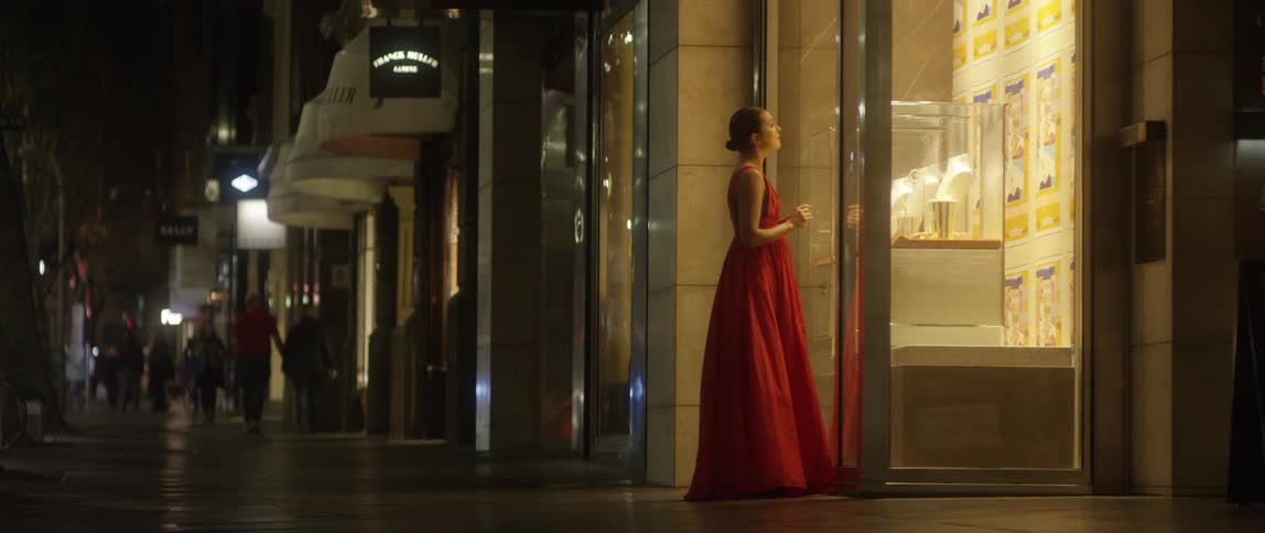 A woman in a sleeveless red ballgown walks to the window display of a jewelry store and looks in